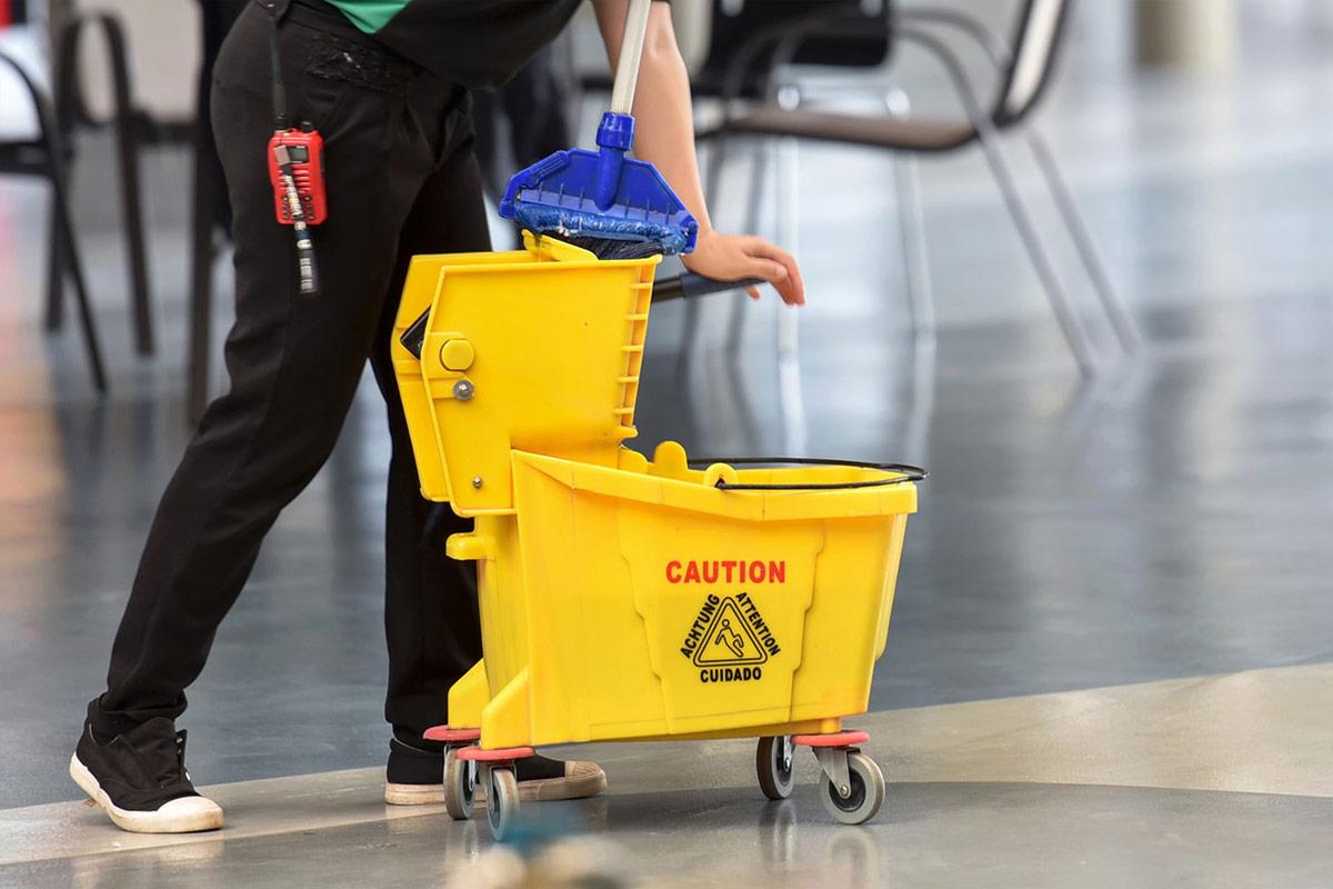 Cleaner rolling a yellow mop bucket across an office floor